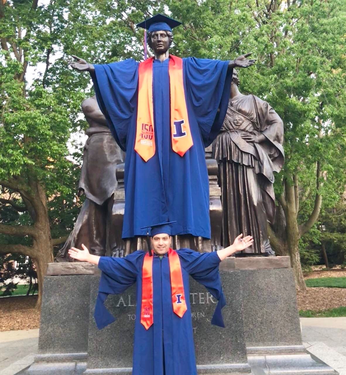 Man in graduation robe and cap poses next to statue