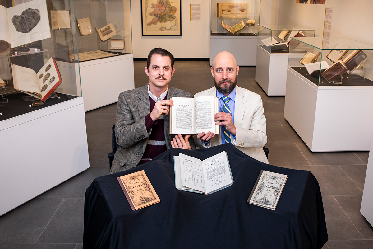 Two men sitting at a table, holding up a book