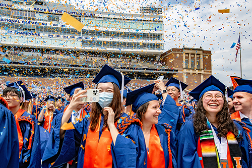 Group of graduates smiling while confetti rains down. Graduate in front snaps a photo on her phone.