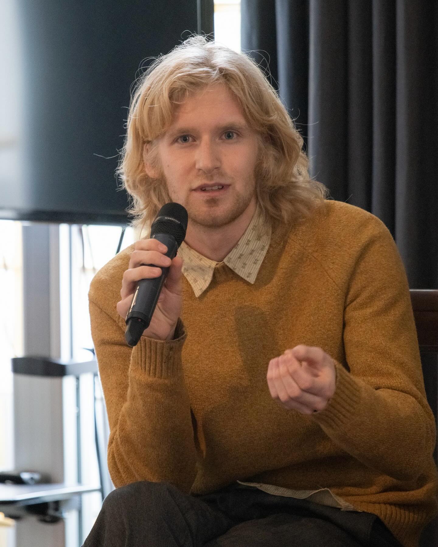 Young man with shoulder length blonde hair in a beige sweater holding a microphone