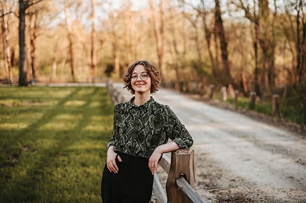 Woman leans on fence with woods behind her 