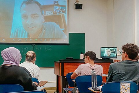 Students sit in a classroom, with a video call projected on a screen in front of them