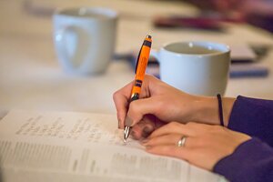 Close-up of female hands with pen writing notes with cups in background.