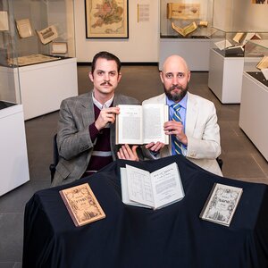 Two men sitting at a table, holding up a book