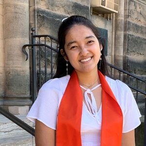 Smiling young woman with dark hair, a white blouse, and an orange graduation stole 