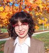 Woman with short dark hair and red glasses poses in front of fall foliage