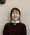 Woman with dark hair and bangs, wearing a brown sweater over a white collared shirt