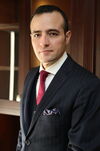 Dark-haired man in dark suit and red tie stands in front of wood background